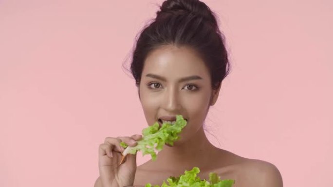 年轻亚洲女性的特写美女肖像，完美光滑健康的皮肤吃沙拉在粉红色的背景下。护肤商业概念。