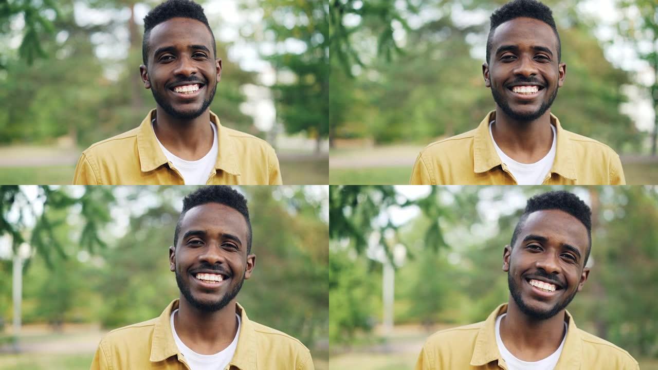 穿着时髦服装的无忧无虑的非洲裔美国人的慢动作肖像站在公园里，独自微笑着看着相机。人和情感概念。