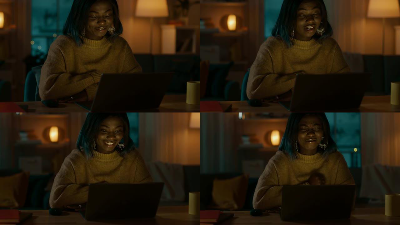 美丽微笑的黑人女孩的肖像坐在她的办公桌前，用笔记本电脑打视频电话。晚上，女孩使用计算机网络摄像头与亲