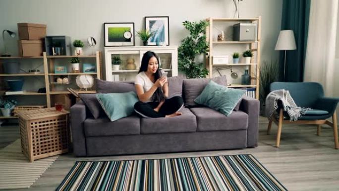 年轻的亚洲妇女正在使用智能手机并在沙发上休息，而机器人真空吸尘器正在用吸尘器代替她。现代技术和家庭概
