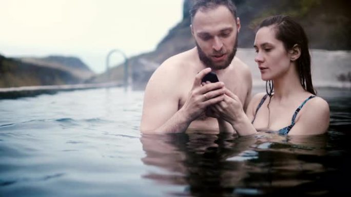 年轻美丽的夫妇在山谷的温泉里游泳，在冰岛一起用智能手机看照片。