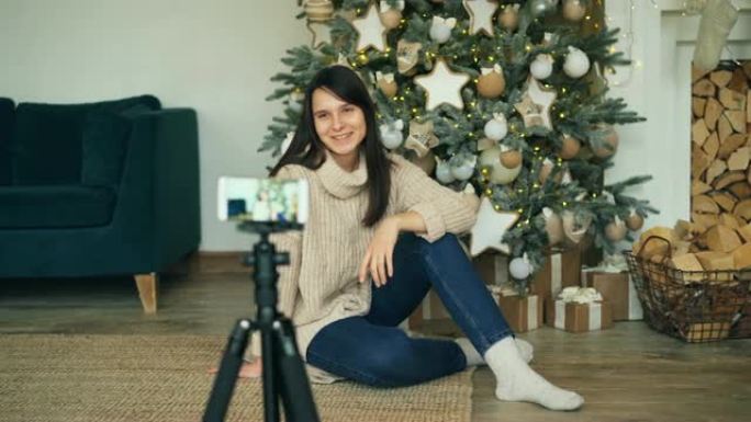 开朗的年轻女士正在录制视频，坐在装饰好的圣诞树附近，看着三脚架上的智能手机相机。女孩正在触摸枞树上的