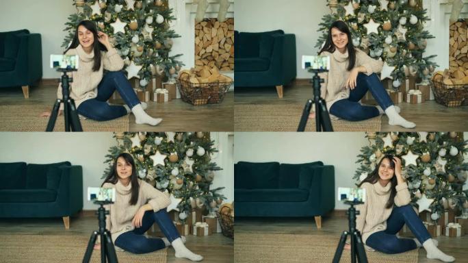 开朗的年轻女士正在录制视频，坐在装饰好的圣诞树附近，看着三脚架上的智能手机相机。女孩正在触摸枞树上的