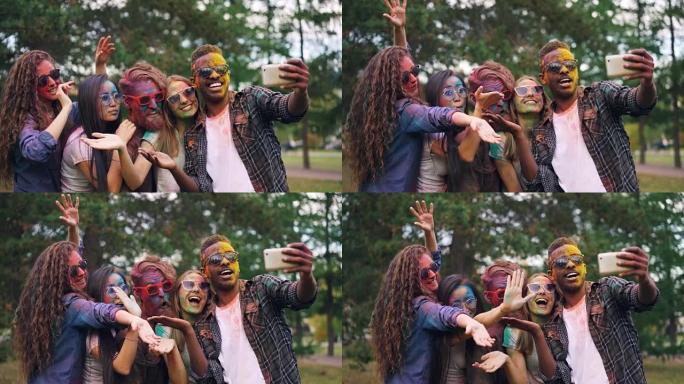 有色面孔的无忧无虑的学生慢动作，用智能手机站在公园的户外并摆姿势拍照。洒红节和照片概念。