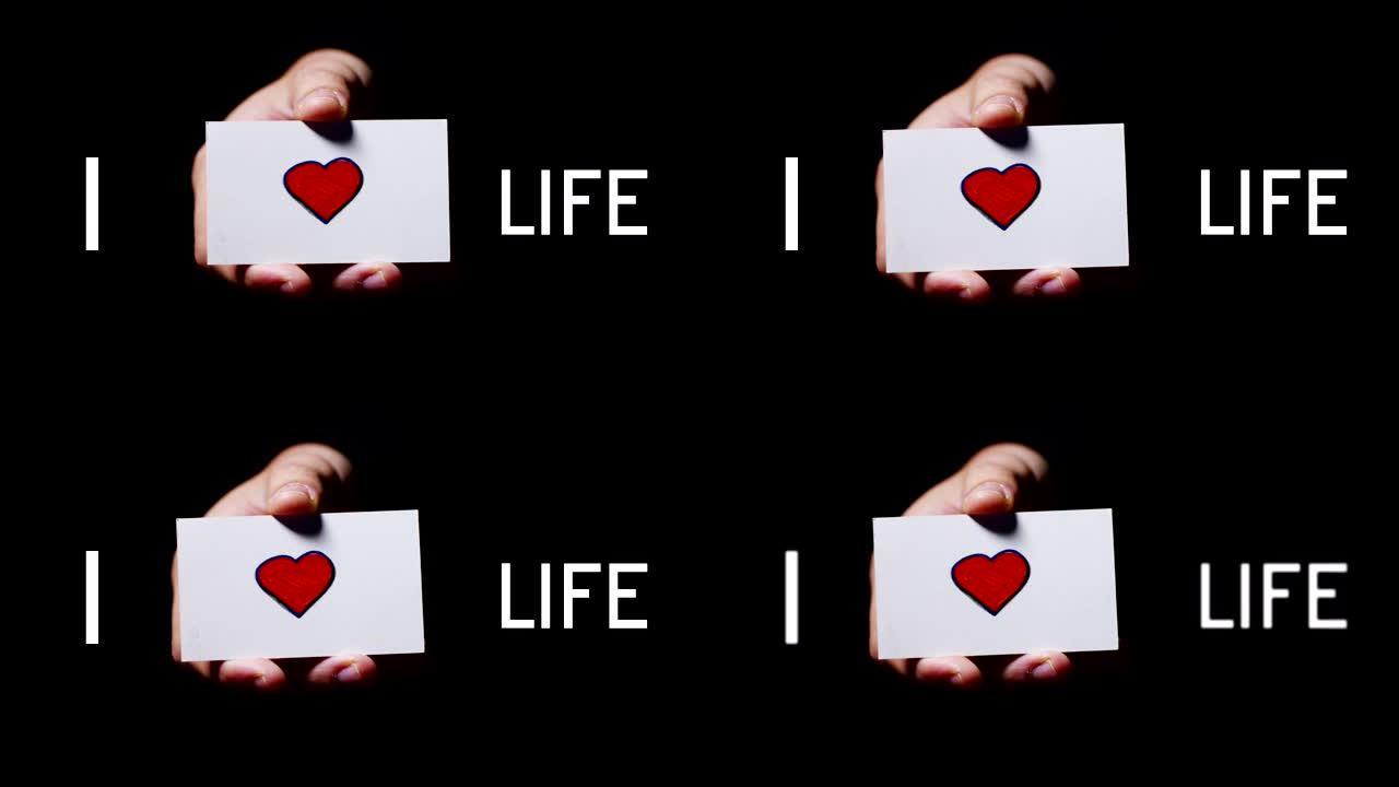一只手展示一张卡片，上面写着: “我喜欢生活”，孤立在黑色背景上。
