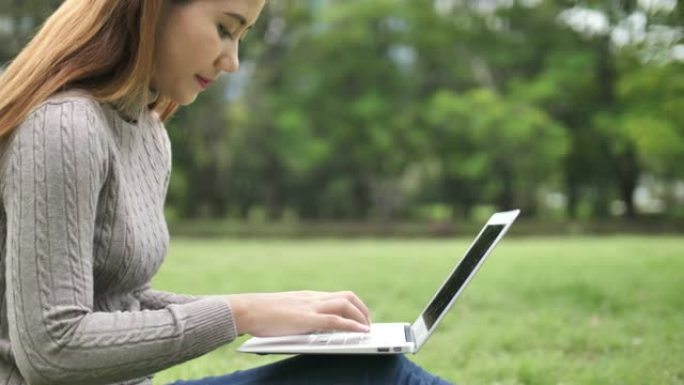 年轻的亚洲妇女在公园里用笔记本电脑打字
