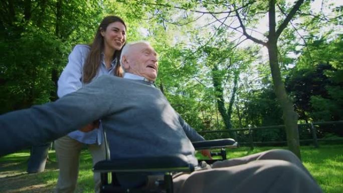 坐在轮椅上的无忧无虑快乐的孙女和祖父的慢动作在阳光明媚的日子里在绿色公园里跑步