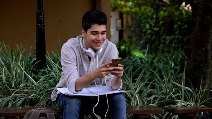 年轻的男学生在智能手机上聊天，脖子上戴着耳机，膝盖上戴着笔记本