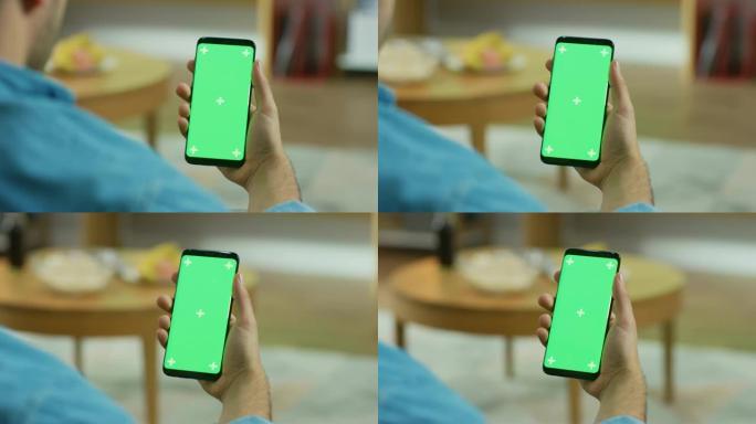 坐在沙发上的男人拿着绿色色度键屏幕触摸屏智能手机。浏览社交网络，阅读新闻或使用应用程序。