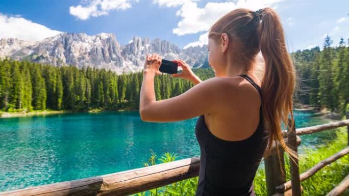 探索多洛米蒂山腰的女游客，站在特伦蒂诺的卡雷扎湖边，背景是拉特马尔山脉，用智能手机拍照