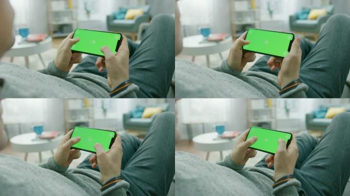 在家中的男人躺在沙发上，使用智能手机，以横向模式水平握住它。按下按钮玩视频游戏。屏幕上有跟踪标记。视
