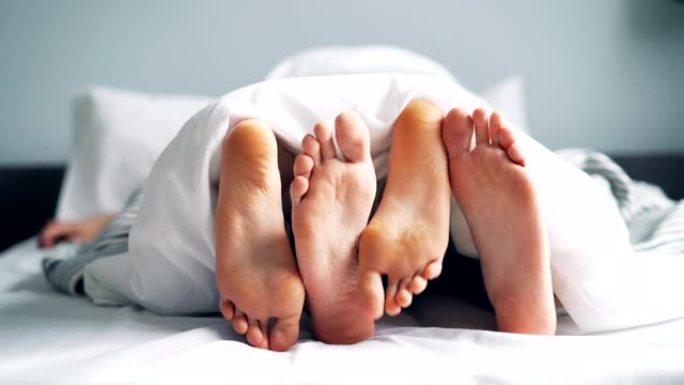女孩和男孩的脚在毯子下的床上爱抚，夫妻享受着关系