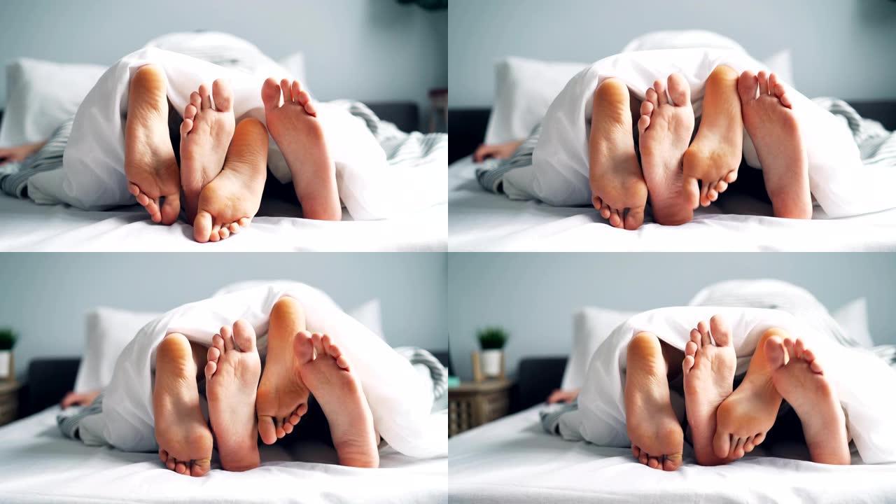 女孩和男孩的脚在毯子下的床上爱抚，夫妻享受着关系