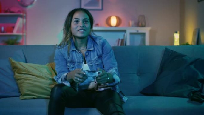 美丽兴奋的年轻黑人游戏玩家女孩坐在沙发上，在游戏机上玩视频游戏并获胜。她玩无线控制器。舒适的房间配有