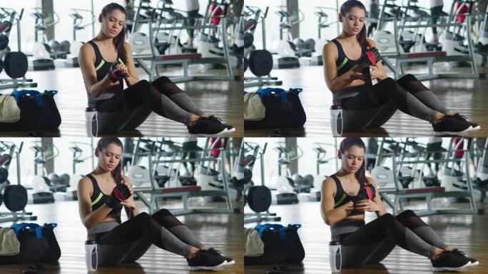 坐在健身房地板上的健康女人用一瓶能量饮料固定鞋子
