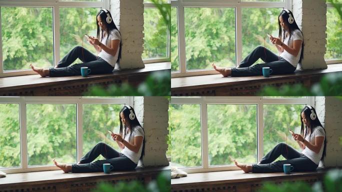可爱的亚洲女孩布鲁内特坐在窗台上，戴着耳机听音乐，用智能手机休息。现代室内、技术和人的概念。