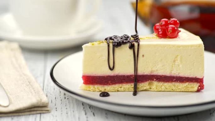 红醋栗芝士蛋糕放在盘子上，上面装饰着红色浆果，上面倒入巧克力釉。背景中模糊的白杯茶和茶壶。滑块镜头，