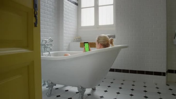 美丽的金发女孩洗澡时使用绿色模拟屏幕智能手机放松。迷人的女孩使用色度键手机浏览社交媒体，看视频，上网