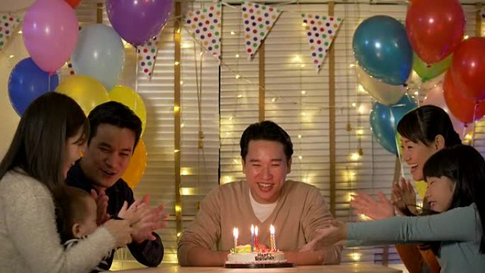 快乐的亚洲男性坐在桌子旁，被家人和朋友包围着，用蛋糕庆祝他的生日。