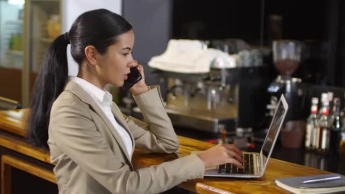 亚洲女商人在酒吧柜台打电话并在笔记本电脑上打字