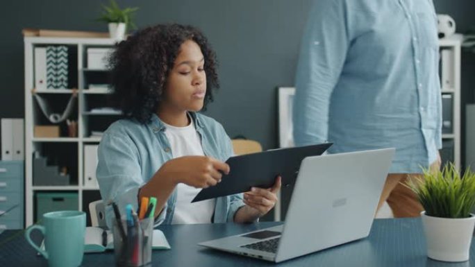 有吸引力的非洲裔美国女士从男同事那里获取文件，然后使用笔记本电脑