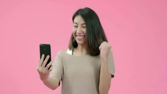 使用智能手机检查社交媒体的年轻亚洲女性在粉红色背景工作室拍摄的休闲服装中感到快乐的微笑。快乐微笑可爱