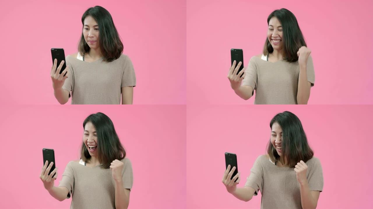 使用智能手机检查社交媒体的年轻亚洲女性在粉红色背景工作室拍摄的休闲服装中感到快乐的微笑。快乐微笑可爱