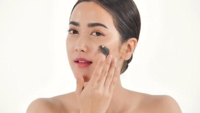 亚洲年轻漂亮的女人在她的颧骨上使用黑色的面霜，在白色背景上使用圆形运动的面霜，皮肤护理，天然化妆品，
