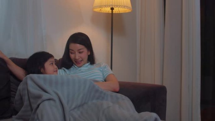 年轻的亚洲家庭和女儿晚上在家看电视。韩国妈妈和小女孩快乐地利用家庭时间躺在客厅的沙发上放松。有趣的妈