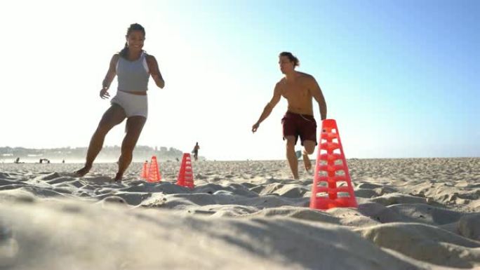 美丽健康的女人在海滩上围绕运动锥训练，私人教练与她平行奔跑