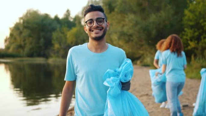 年轻的阿拉伯志愿者带着垃圾袋在湖岸微笑，背景人物