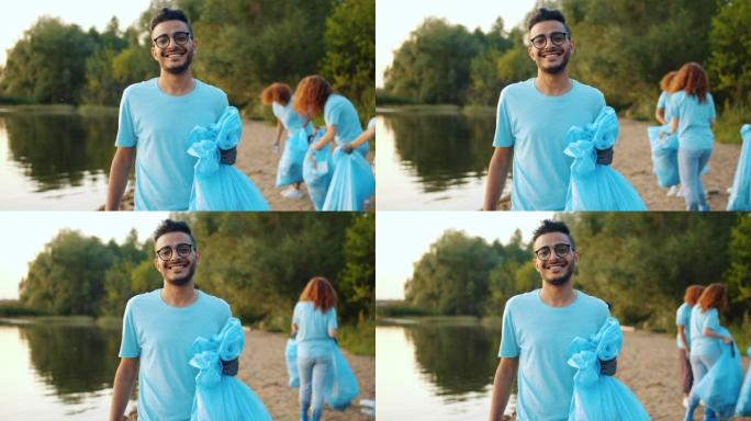 年轻的阿拉伯志愿者带着垃圾袋在湖岸微笑，背景人物