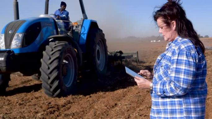 女性农民使用数字平板电脑并监视拖拉机在大型农场上耕作农田