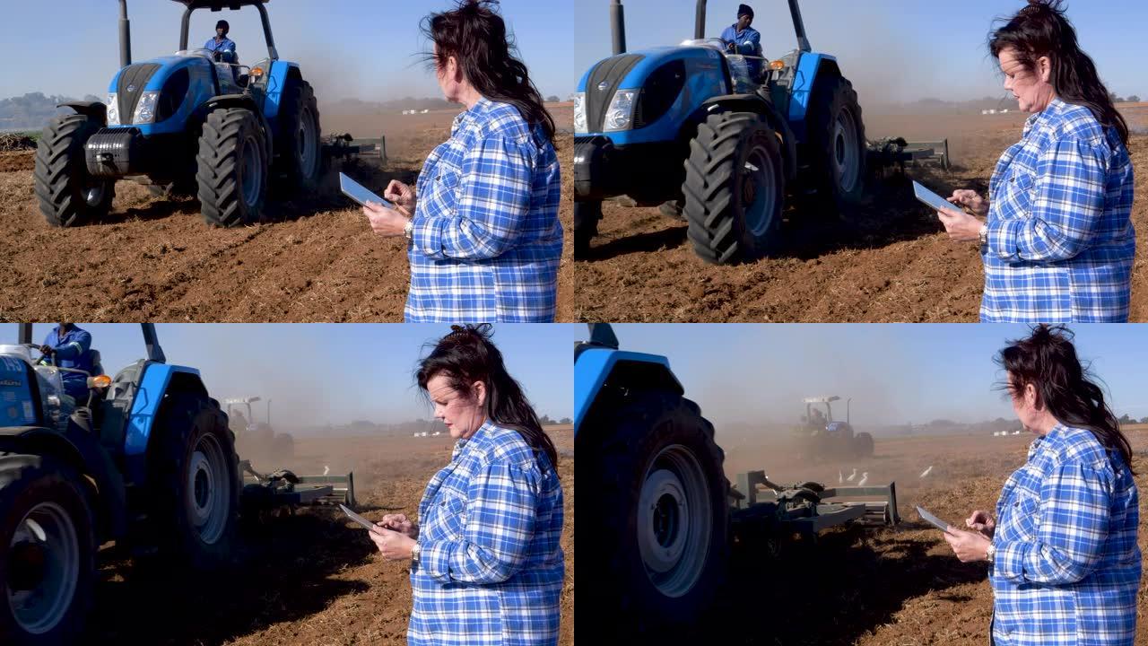 女性农民使用数字平板电脑并监视拖拉机在大型农场上耕作农田