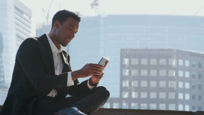 千禧年黑人商人穿着西装和白衬衫坐在泰晤士河堤岸上，用他的智能手机特写