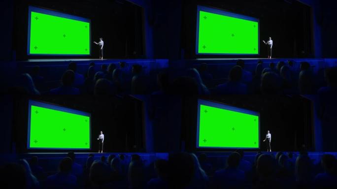 鼓舞人心的演讲者在电影院后面用绿屏，模型，色键向观众展示新产品。女首席执行官在业务现场活动或设备展示