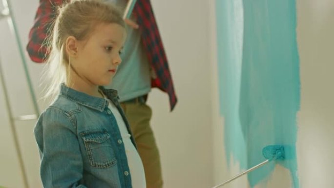 穿着牛仔裤外套的可爱的小女孩正在粉刷墙壁。她用覆盖着浅蓝色油漆的滚筒绘画。父亲和女儿一起画画。家里的