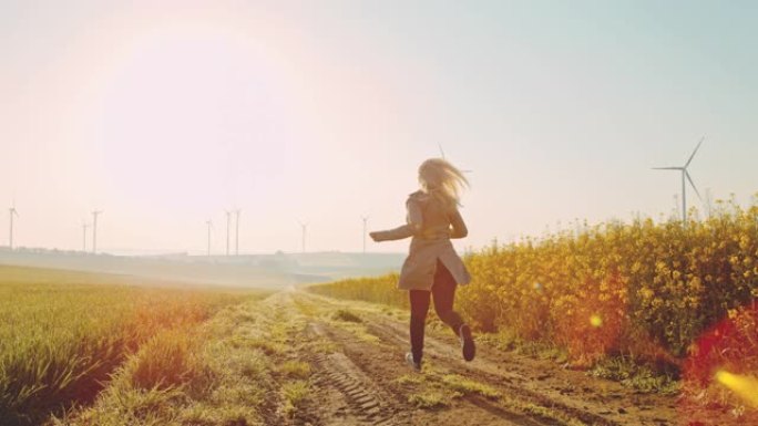 SLO MO女人沿着油菜籽田野奔跑，远处有风力涡轮机