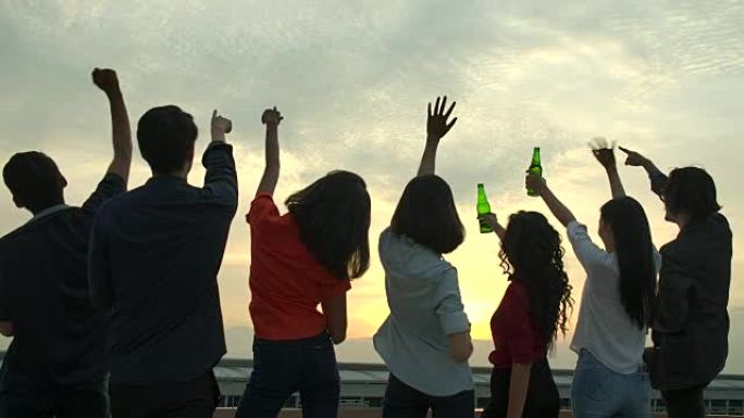 一群年轻的朋友跳舞，玩得开心，在日落背景的夏季屋顶派对上，用烤面包和叮当响的举杯庆祝。慢动作。