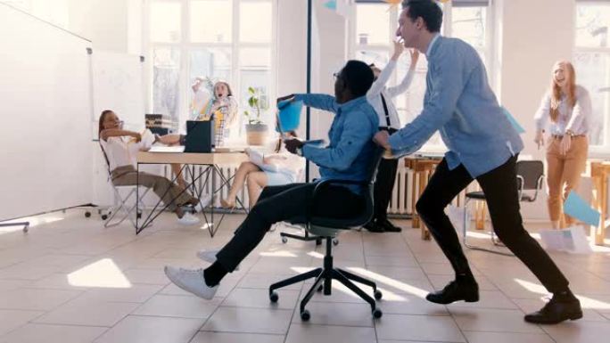 慢动作疯狂有趣的年轻黑人商人骑着办公椅，用团队在空中扔纸庆祝成功。