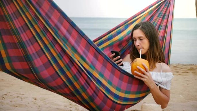 严肃迷人的年轻自由职业者女人躺在海洋海岸的海滩吊床上，水果鸡尾酒使用智能手机应用程序。