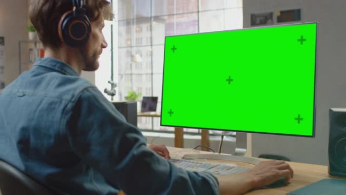 年轻的专业创意员工在他的个人计算机上工作，并带有绿色大屏幕模拟显示屏。他在一个很酷的办公室阁楼里工作