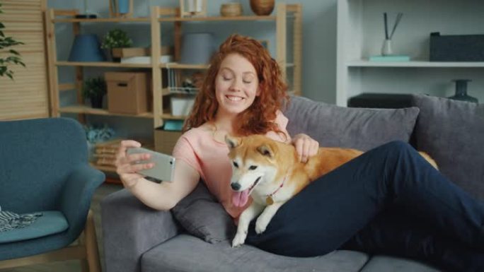 快乐的女孩在沙发上用智能手机摄像头带着可爱的狗
