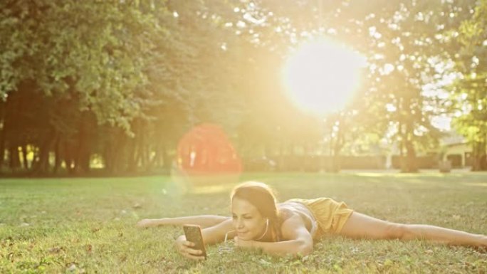 女士灵活的年轻女子练习劈叉，在阳光明媚的公园使用智能手机