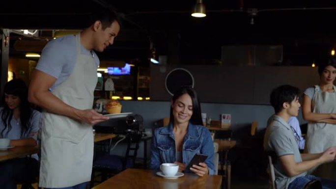 友好的服务员为在智能手机上聊天并在面包店喝咖啡的女顾客带来松饼