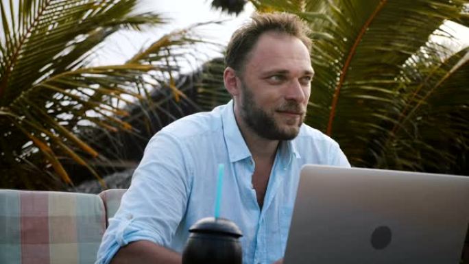 快乐微笑的高加索商人使用笔记本电脑的特写镜头，在异国情调的棕榈树下寻找创意灵感