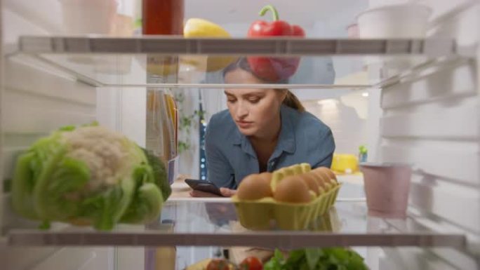 年轻女子打开冰箱门，在智能手机中检查食谱，然后取出一盒鸡蛋。准备健康餐的女人。充满健康食物的厨房冰箱