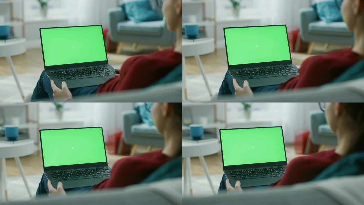 家里的年轻女子在带有绿色模拟屏幕的笔记本电脑上工作。她坐在他舒适客厅的沙发上。肩膀上的相机拍摄