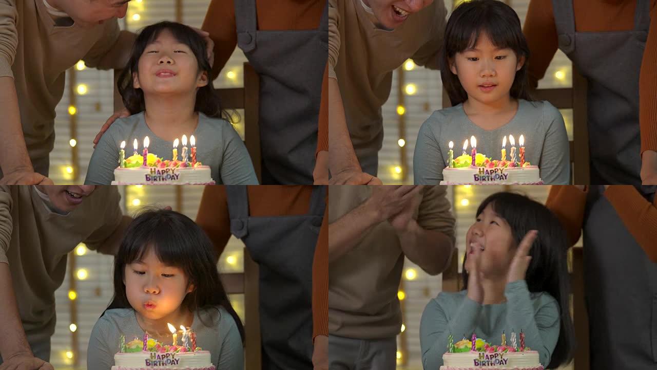 一个可爱的亚洲小女孩坐在餐桌旁，在生日蛋糕上吹蜡烛，而她的家人站在后面，为她唱歌。她露出灿烂的笑容，