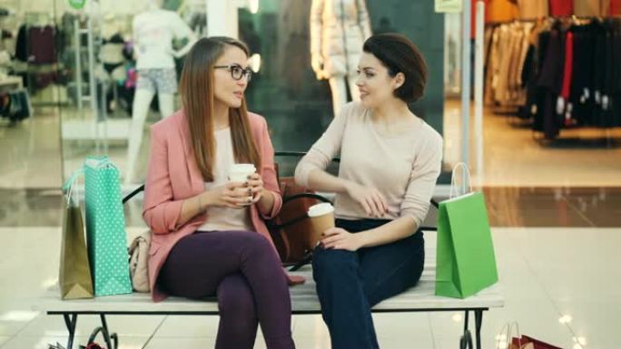 漂亮的年轻女子在商场里聊天，拿着外卖饮料坐在长椅上，纸袋站在旁边。青年生活方式和商店概念。
